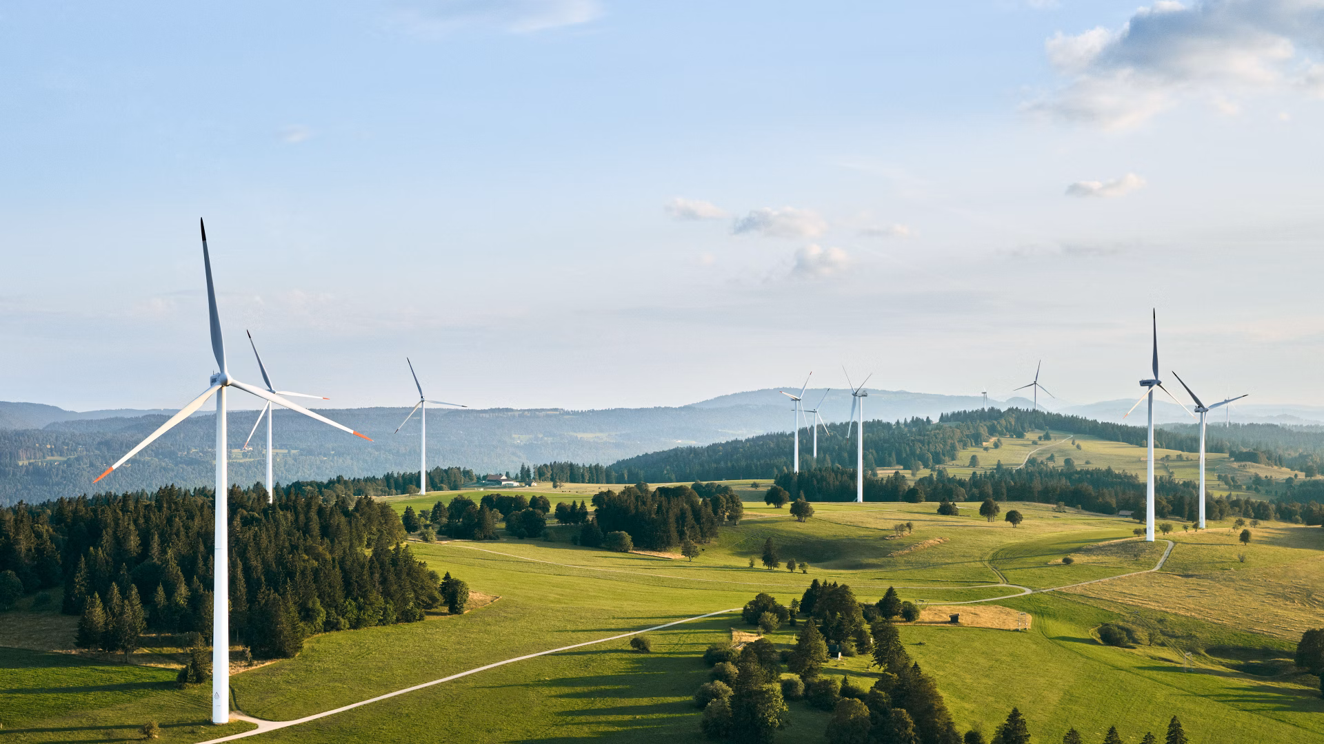 Windturbinen auf einer grünen Hügellandschaft mit Wäldern und Wegen.