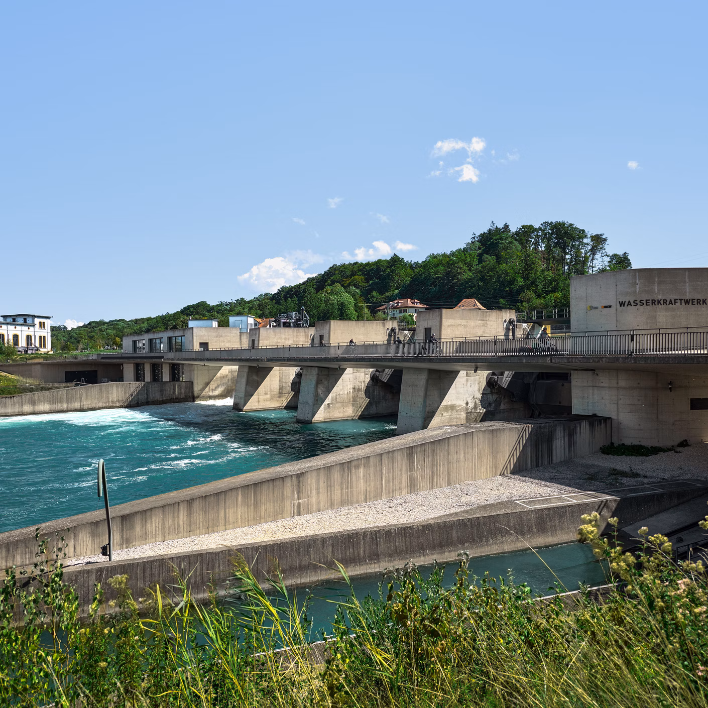 Wasserkraftwerk mit Wehr über einem türkisfarbenen Fluss unter blauem Himmel
