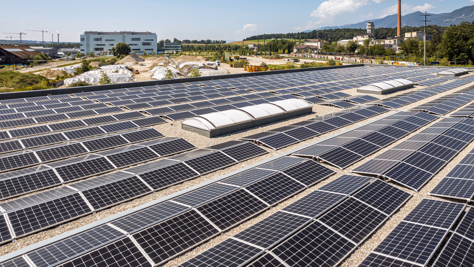 Solaranlage auf einem grossen Flachdach in einem Industriegebiet mit Gebäuden und Hügeln im Hintergrund