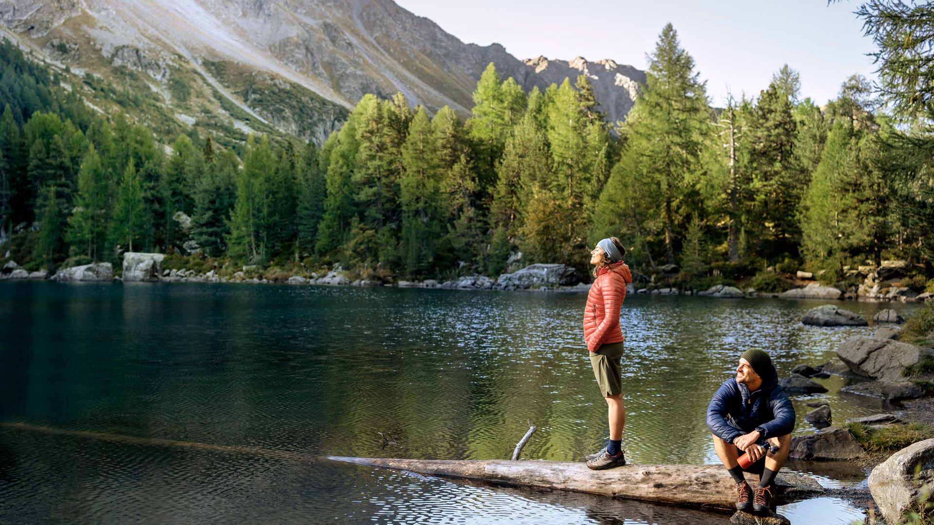 Zwei Personen an einem Bergsee mit Wald und felsigen Gipfeln im Hintergrund