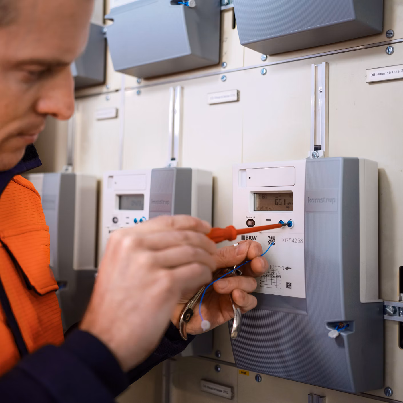 Techniker in orangefarbener Arbeitskleidung prüft Messgeräte in einem Schaltschrank.