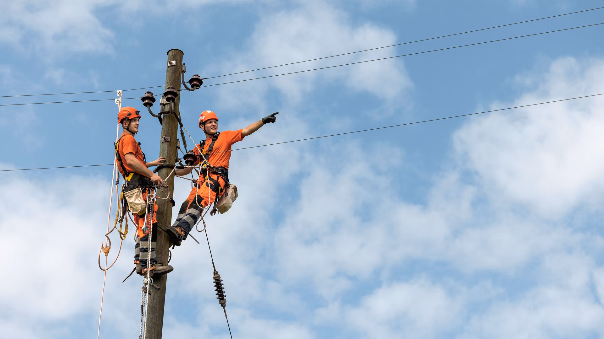 Zwei Arbeiter mit Sicherheitsausrüstung auf einem Strommast vor blauem Himmel mit Stromleitungen