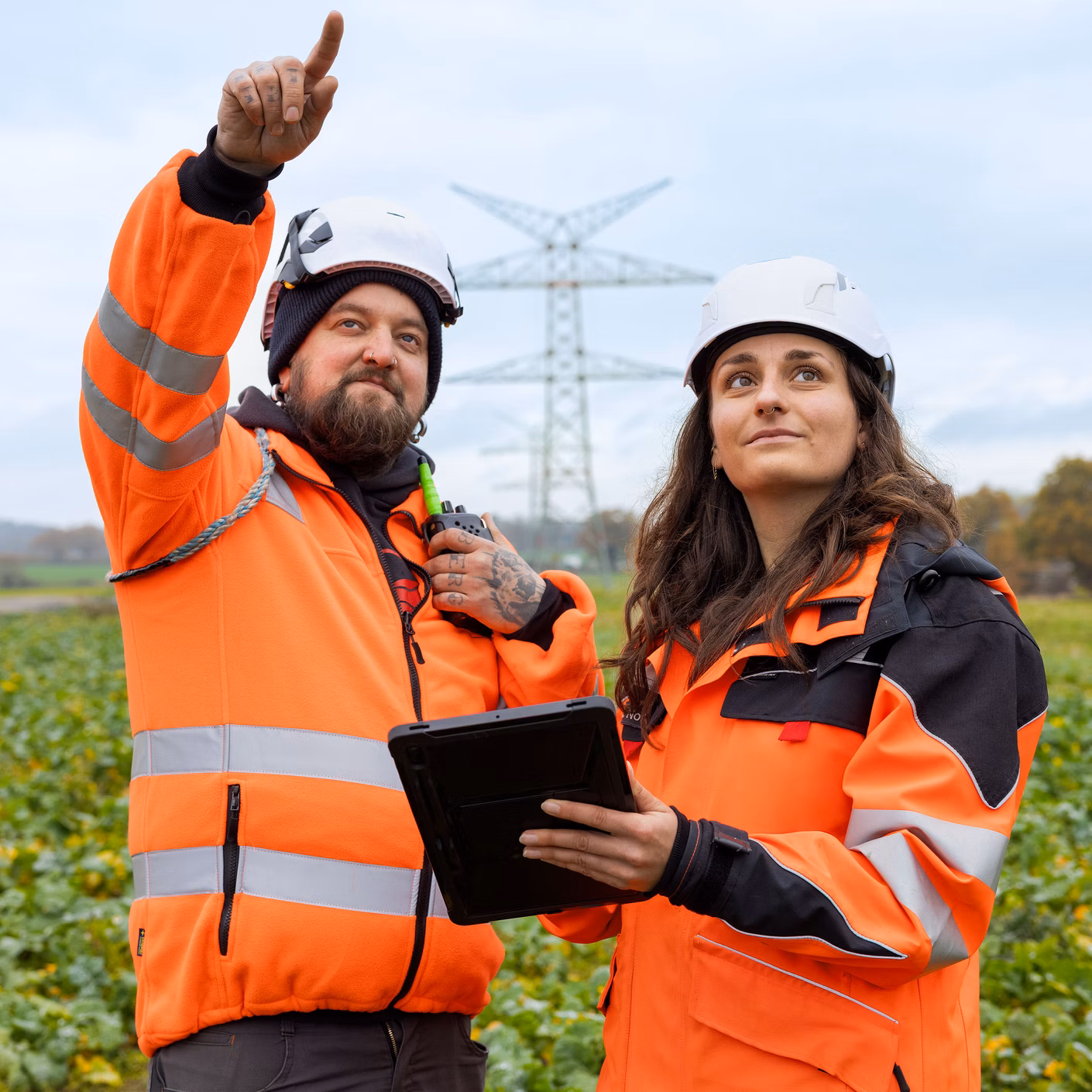 Zwei Personen in oranger Warnkleidung und Helmen stehen auf einem Feld vor einem Strommast; eine Person zeigt in die Ferne.
